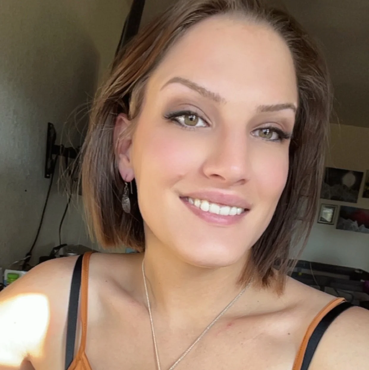 A woman with chin-length brown hair, green eyes, and light makeup, smiling indoors. She is wearing a black and orange spaghetti strap top, a silver necklace, and earrings.