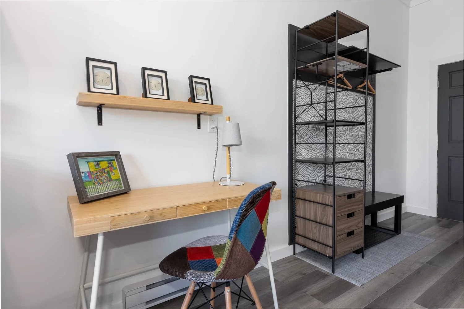 Home office with a wooden desk, colorful patchwork chair, floating shelf with framed art, and a black metal storage unit with wood drawers, patterned wallpaper, and gray flooring.