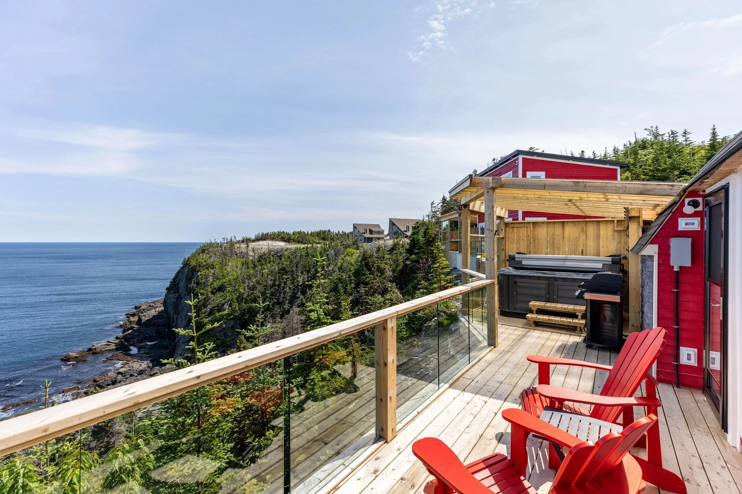 Balcony with red Adirondack chairs overlooking cliffs and ocean with houses on hillside and forest in the background.