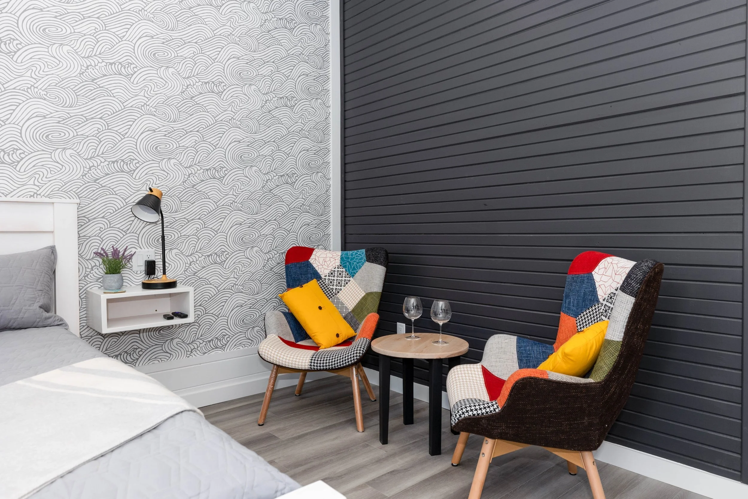 A cozy bedroom corner with a bed, a white nightstand holding a potted plant and a black and gold desk lamp, and two patterned chairs with colorful pillows beside a small round table with two wine glasses. The wall behind features a white and black wave pattern on the left and black horizontal slats on the right.