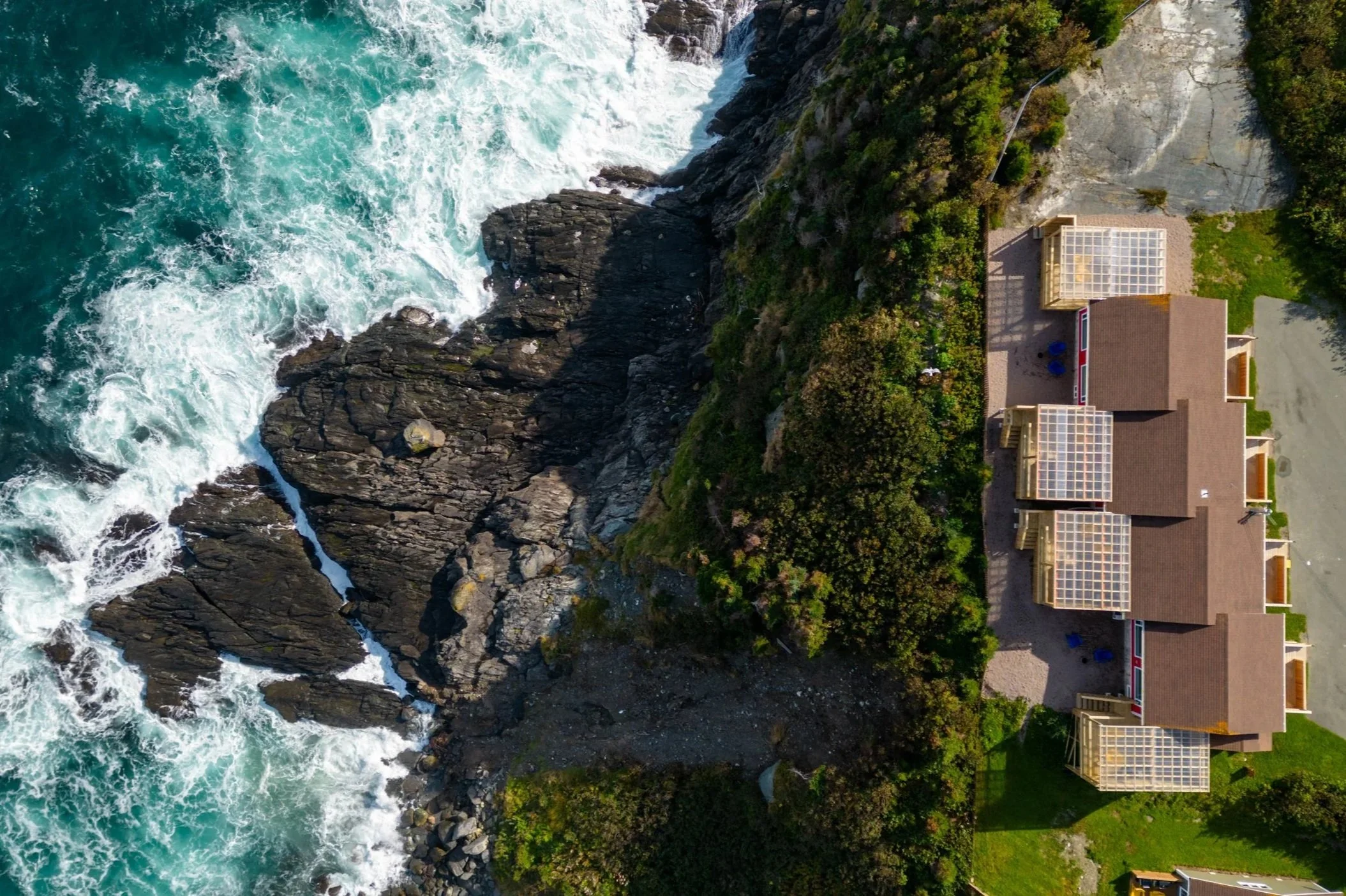 Aerial view of a house along the rocky edge of the ocean, with blue-green water crashing against the rocks.