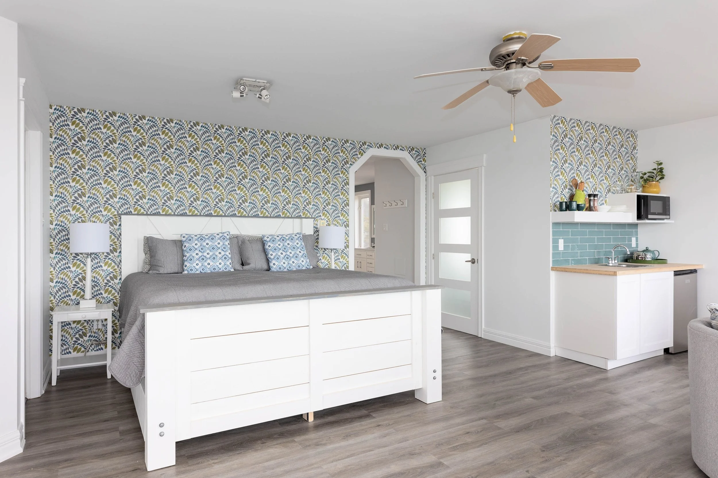 A bedroom with a white bed frame, grey bedding, and blue patterned pillows. The wall behind the bed has blue, green, and beige patterned wallpaper. There are white nightstands with white lamps on either side of the bed. The room has wooden floors and a ceiling fan. To the right, part of a kitchen area is visible with a white counter, teal subway tile backsplash, a microwave, and a potted plant.