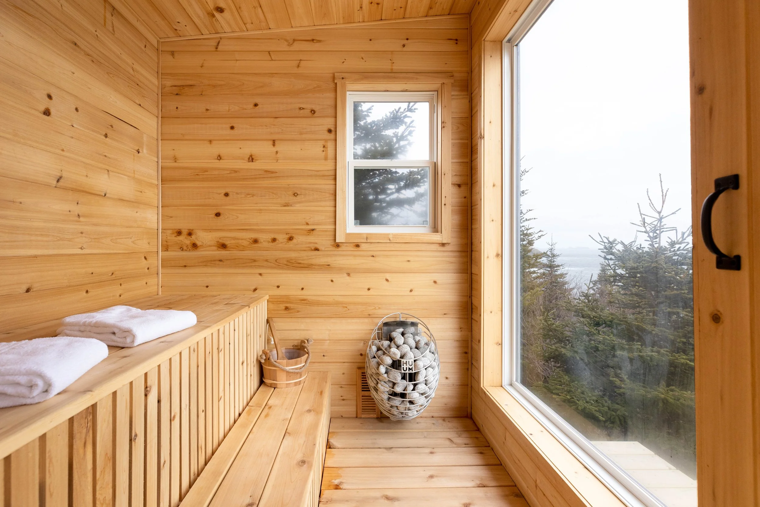 Interior of a wood-paneled sauna with towels, a basket of stones, and large windows showing trees outside.