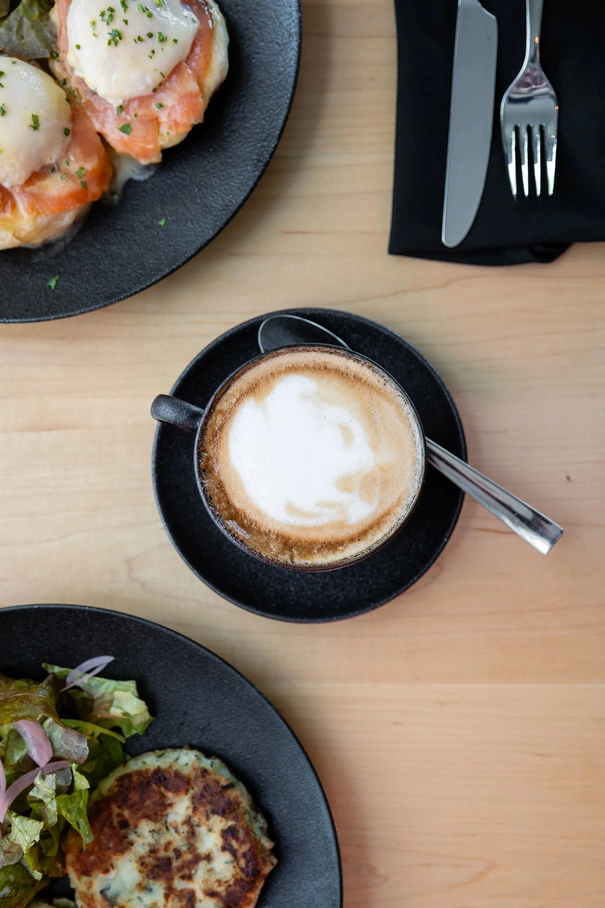 A cup of cappuccino with foamed milk on top, served on a black saucer. Part of a plate with food, including a salad and a grilled dish, is visible in the corner on a wooden table.