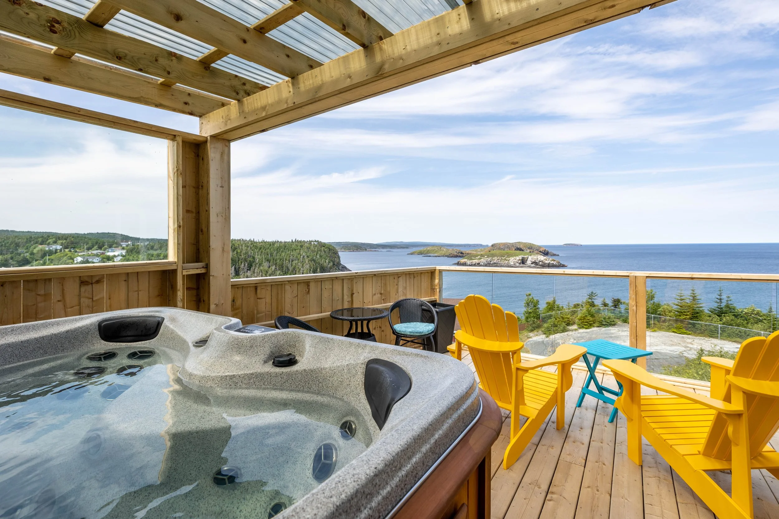 A balcony overlooking a coastal landscape with a hot tub in the foreground, surrounded by colorful chairs and outdoor furniture, with a view of water, small islands, and greenery under a partly cloudy sky.