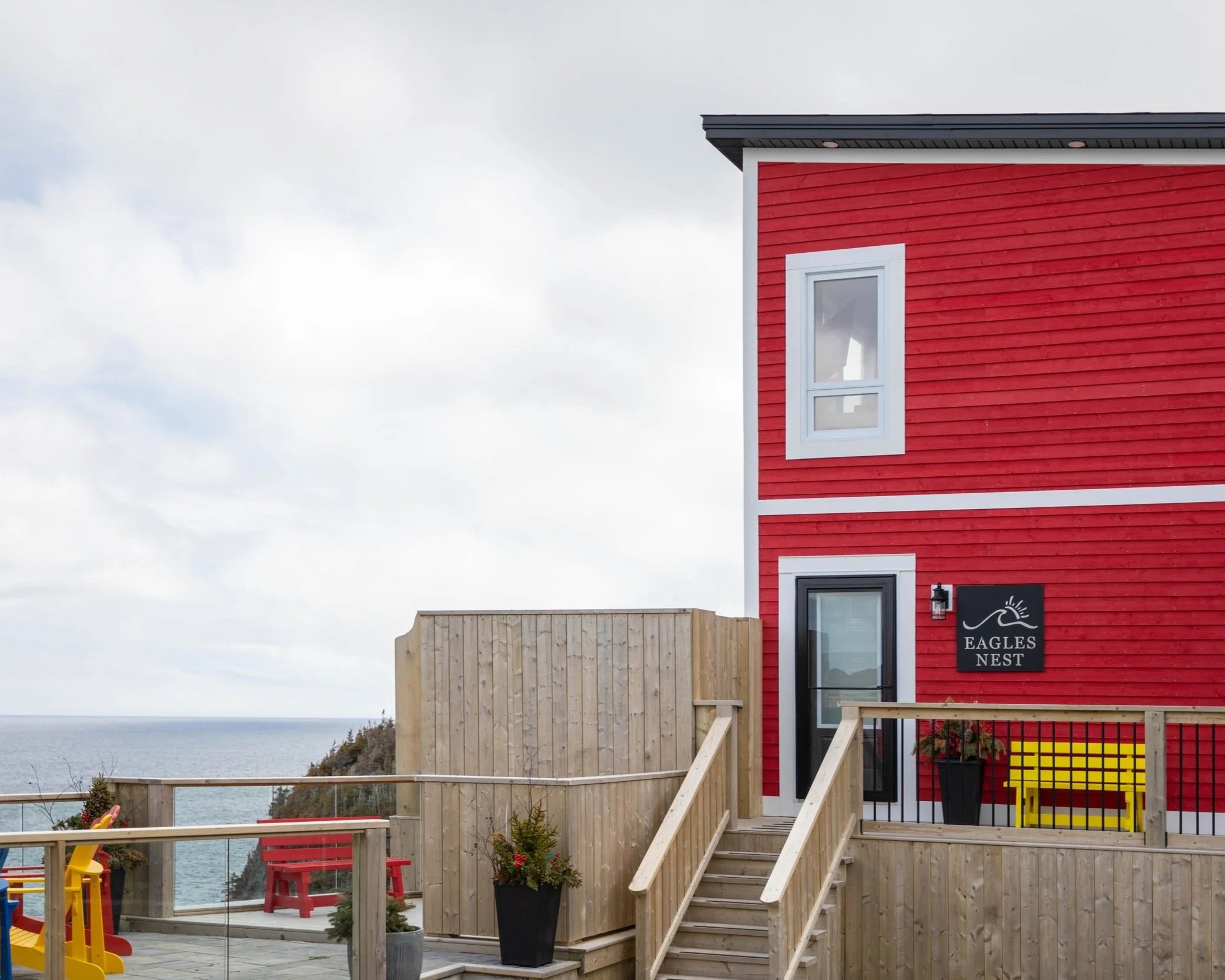 Red wooden building with white-framed windows and black sign reading "Eagles Nest" on a deck with stairs, overlooking a body of water and cloudy sky.