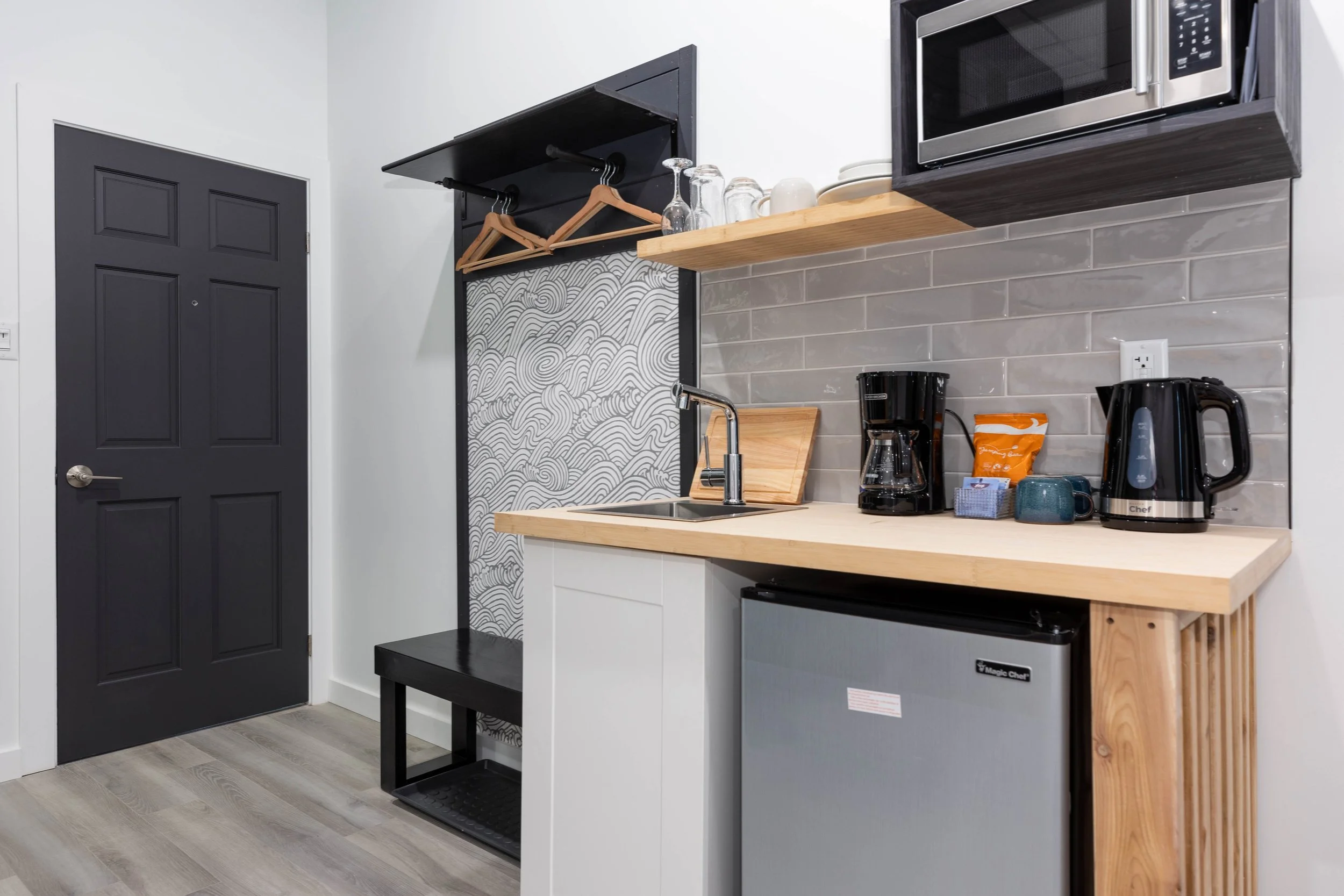 A small kitchenette with a dark door, white walls, and light wood flooring. The kitchen area has a gray and white tile backsplash, a soap dispenser, a black coffee maker, an electric kettle, a microwave oven, and a small mini refrigerator. There are minimalist open shelves holding glasses, cups, and plates, with a black wall-mounted rack with wooden hangers for hanging clothes.