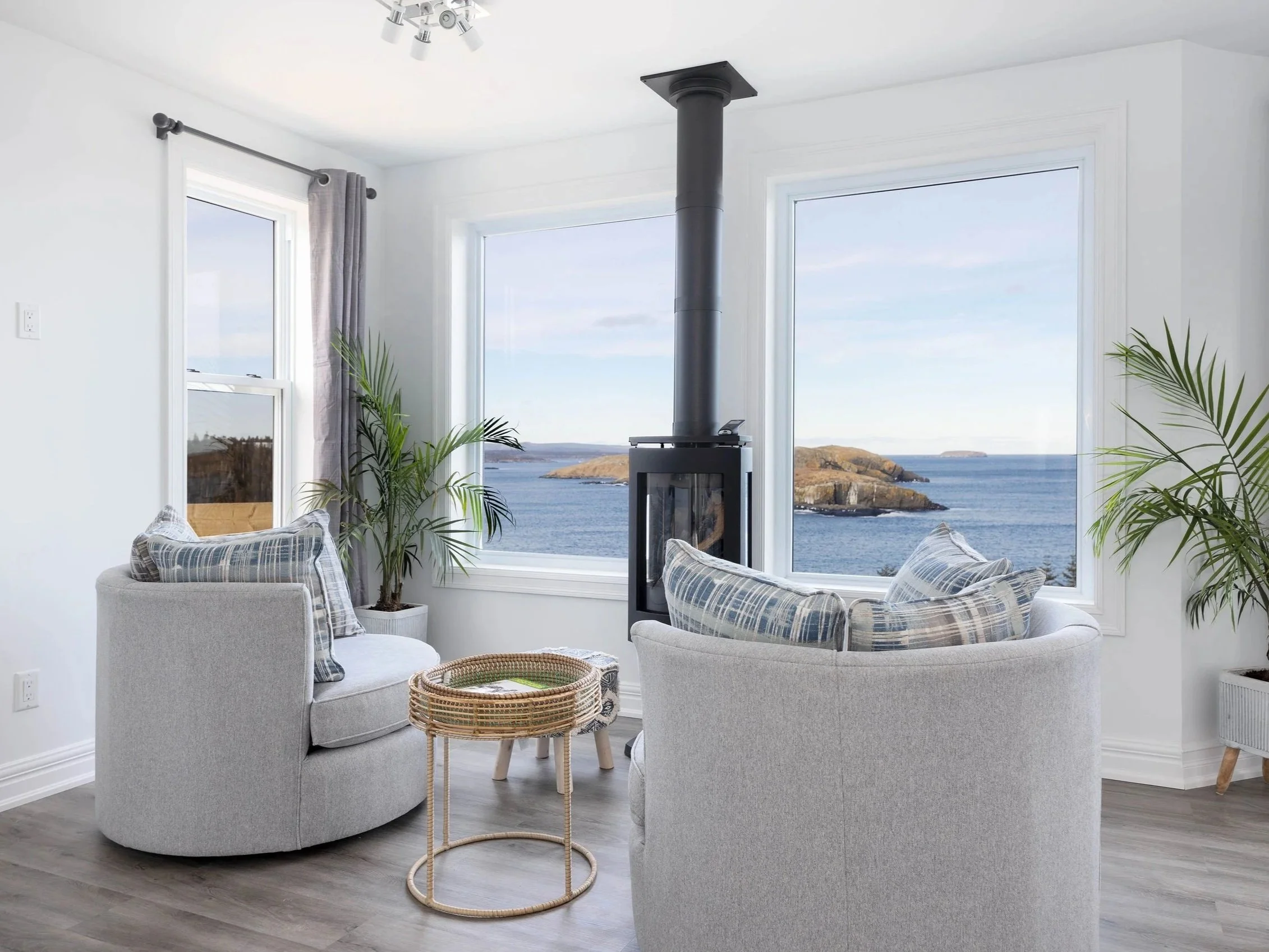 A living room with large windows showing a view of the ocean and rocky islands. There are two light gray sofas with plaid cushions, a small wicker side table, and indoor plants. A gray curtain hangs by a smaller window, and a modern ceiling light fixture is on the ceiling.