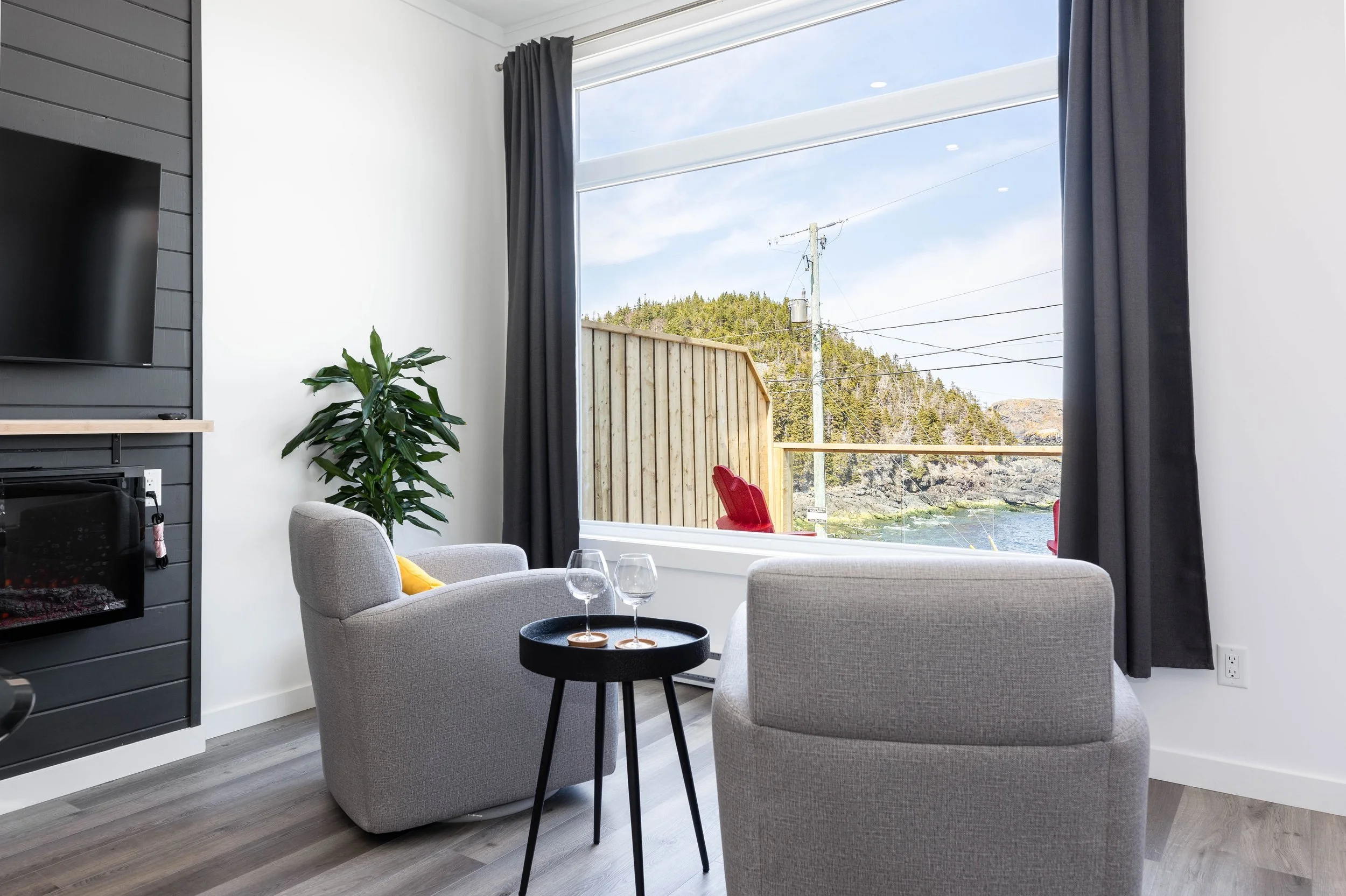 Living room with two light gray armchairs and a small black side table with two empty wine glasses, a plant near the window, and a window with black curtains showing a view of trees, power lines, and a body of water outside.