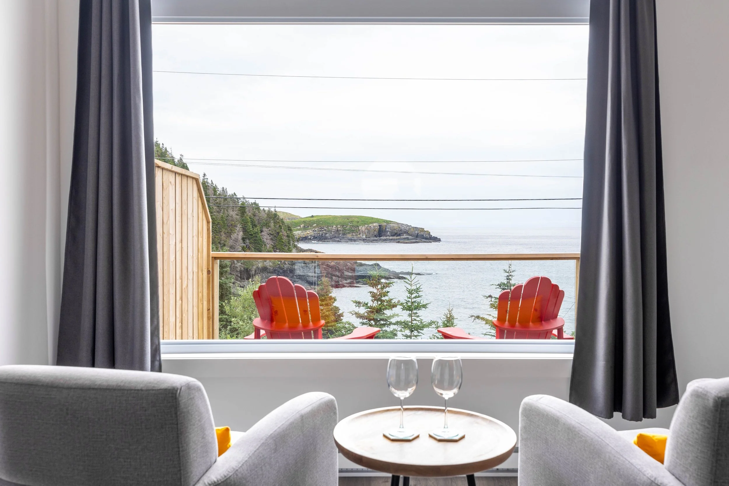 Living room with a view of an outdoor balcony overlooking a coastal landscape, with red chairs on the balcony, framed by dark drapes, and glass of champagne on a table inside.