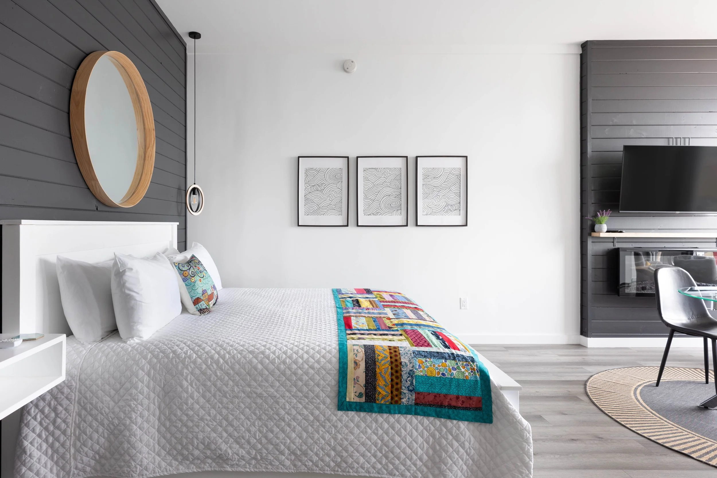A bedroom with a white bed, colorful quilt, black and white framed wall art, flat-screen TV, gray and white walls, and a round striped rug.