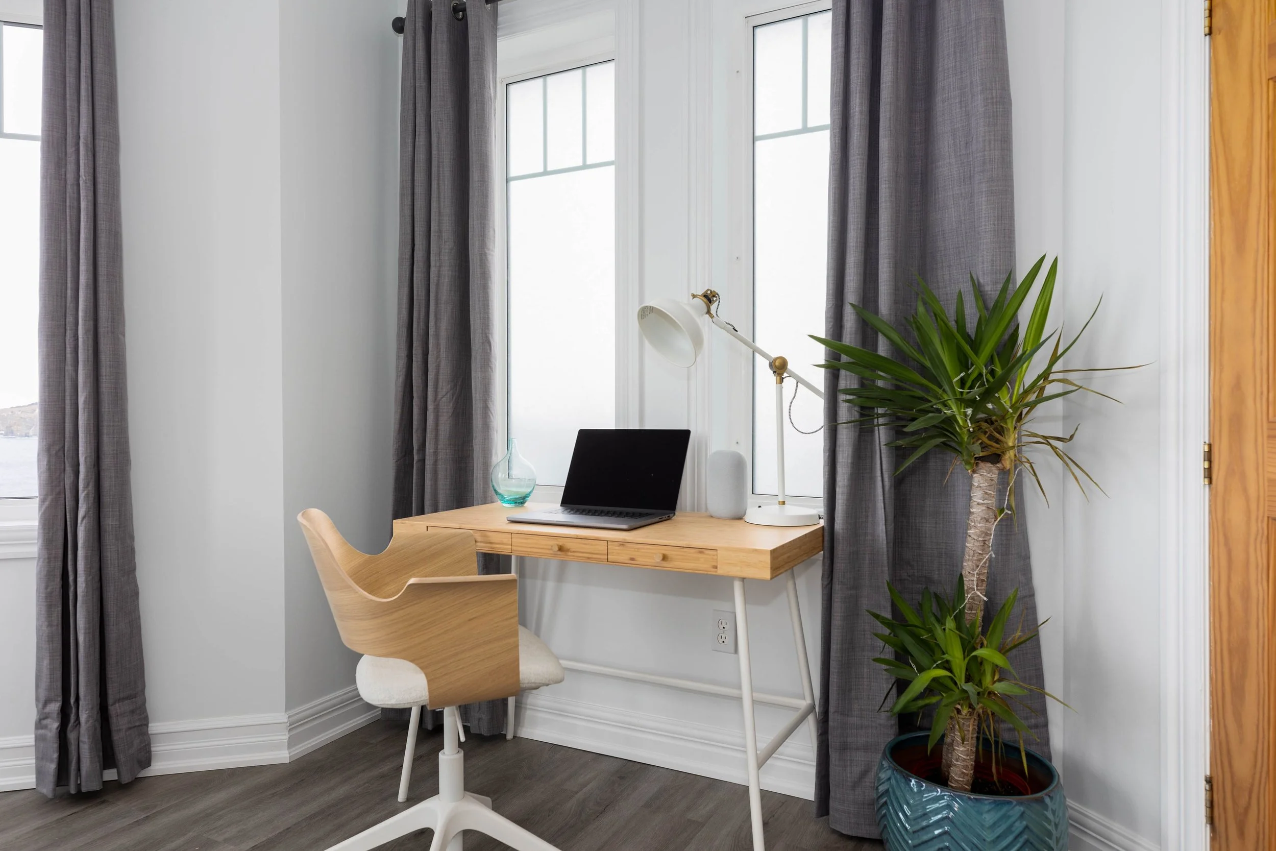 A home office corner with a wooden desk holding a laptop, a transparent glass vase, and a white adjustable desk lamp. A modern wooden chair with a white cushioned seat is in front of the desk. The space features large windows with gray curtains and a tall potted plant with long green leaves in a decorative blue pot. The floor is dark wood, and part of a wooden door frame is visible on the right.