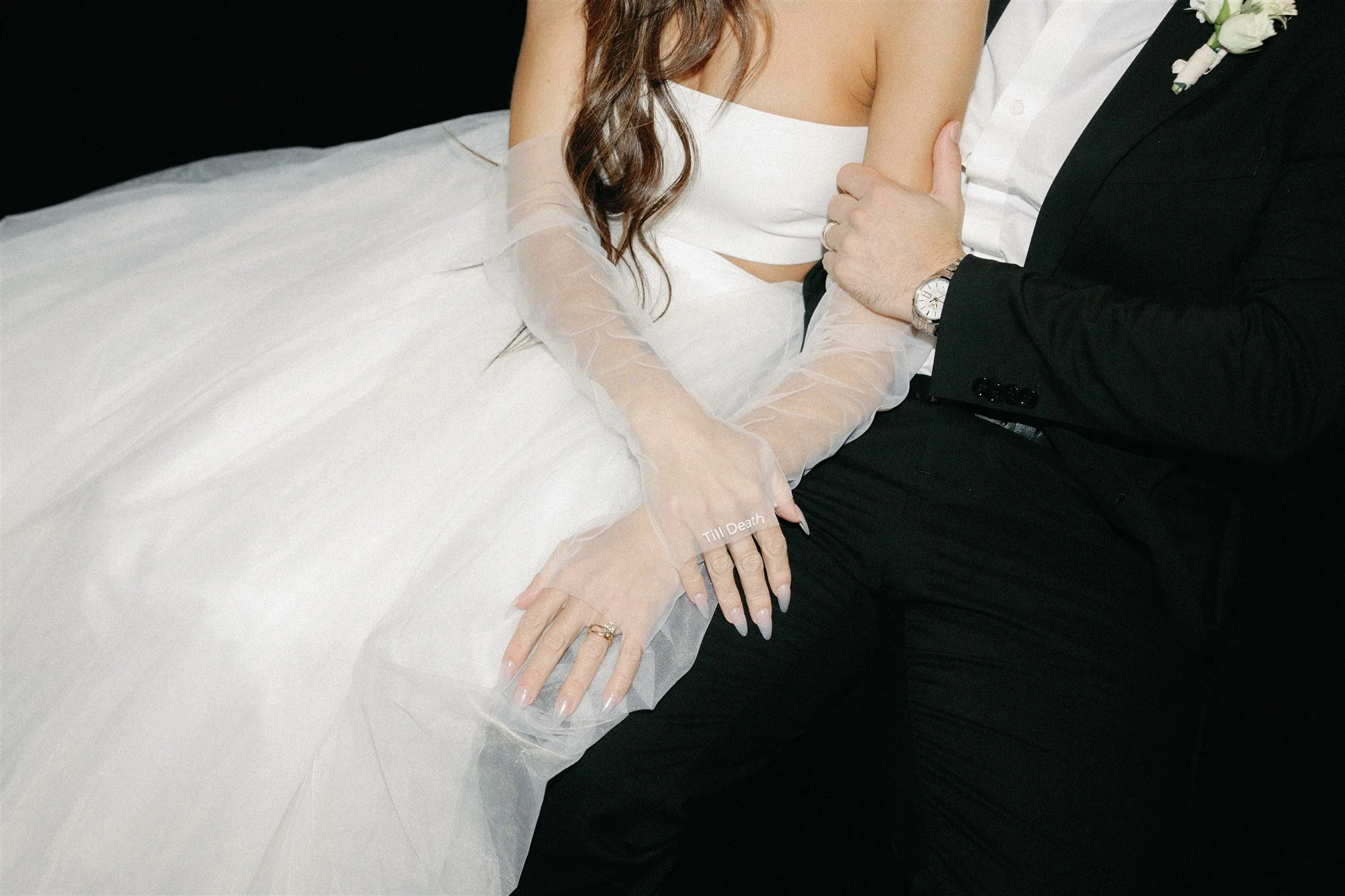 Close-up of a bride and groom sitting together, with the bride dressed in a white wedding gown and the groom in a black suit. The bride's left hand, decorated with rings, rests on her lap, touching the groom's arm, and she wears a PVC glove with the text 'Till Death' on it.