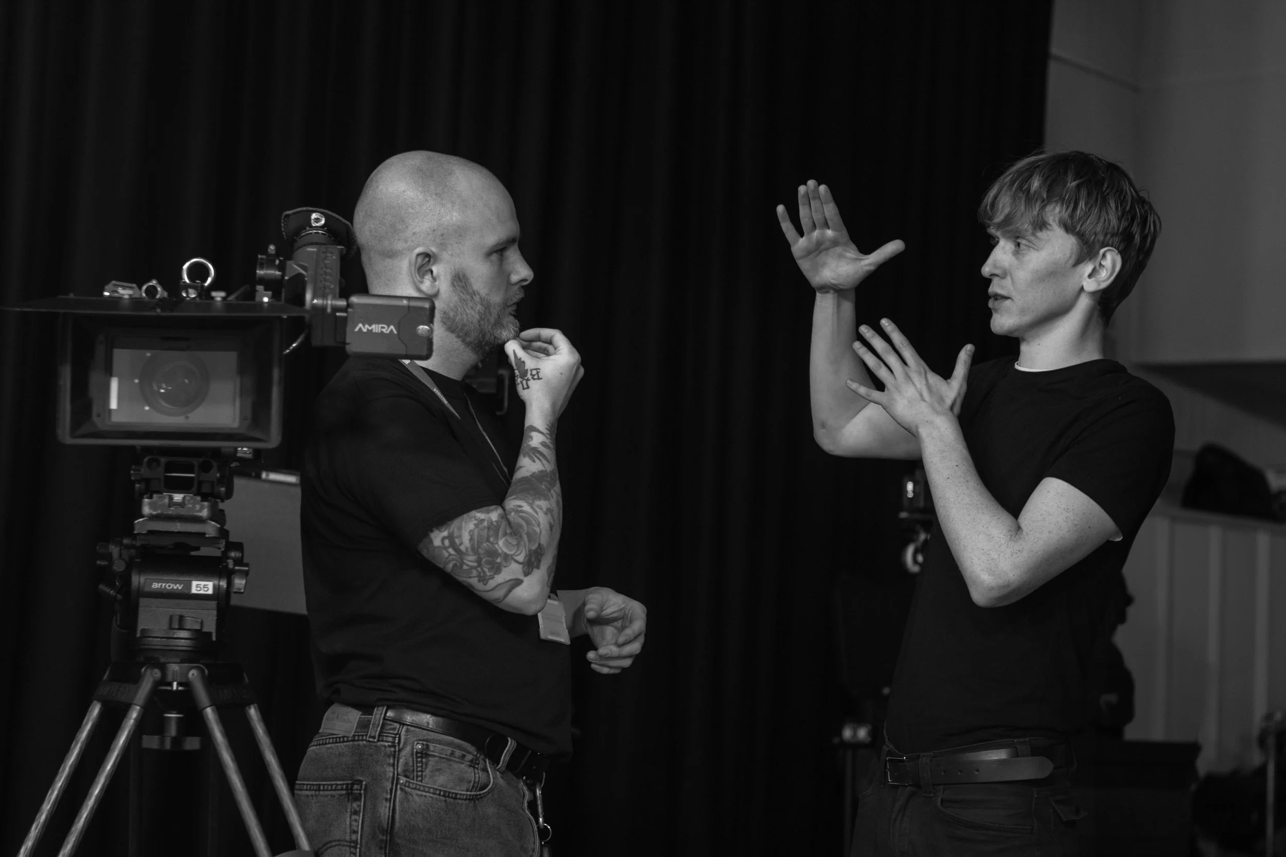 Two filmmakers on set; one gestures to frame a shot with hands, while the other listens beside a large camera.