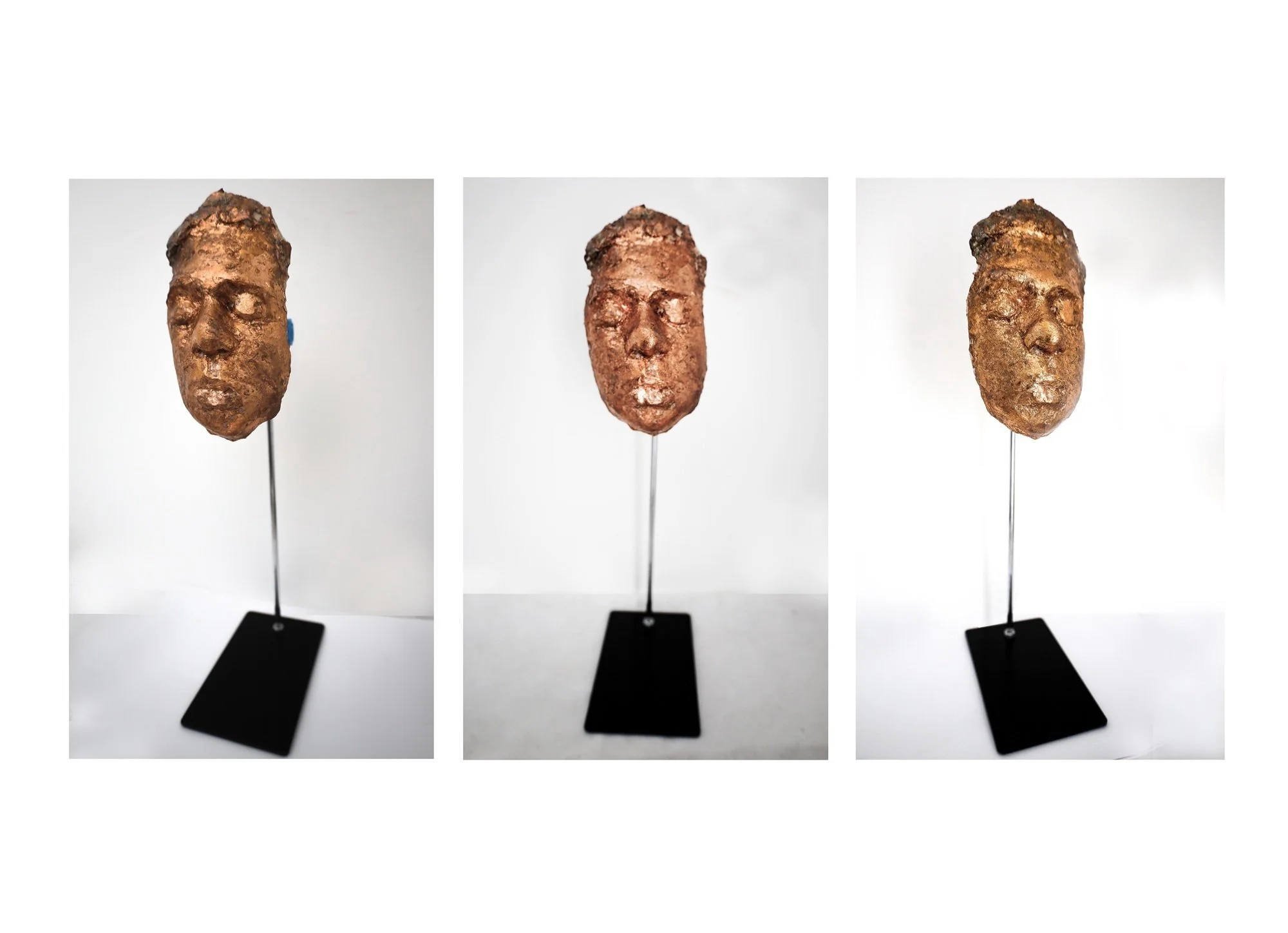 Three sculpted metal masks mounted on black stands, each depicting a human face with distinct features and textures, set against a white background.