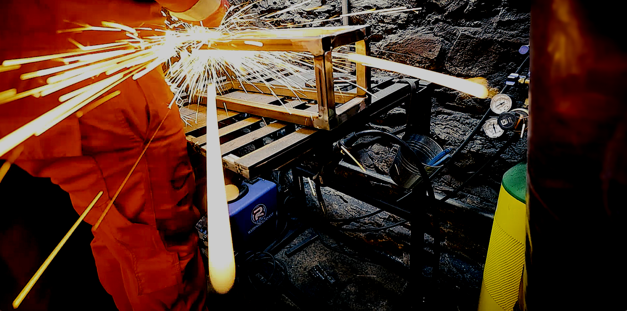 A person welding metal, creating bright sparks, on a workshop space with tools and equipment nearby. The image was taken at the Welding Space Studio, Plymouth, UK.