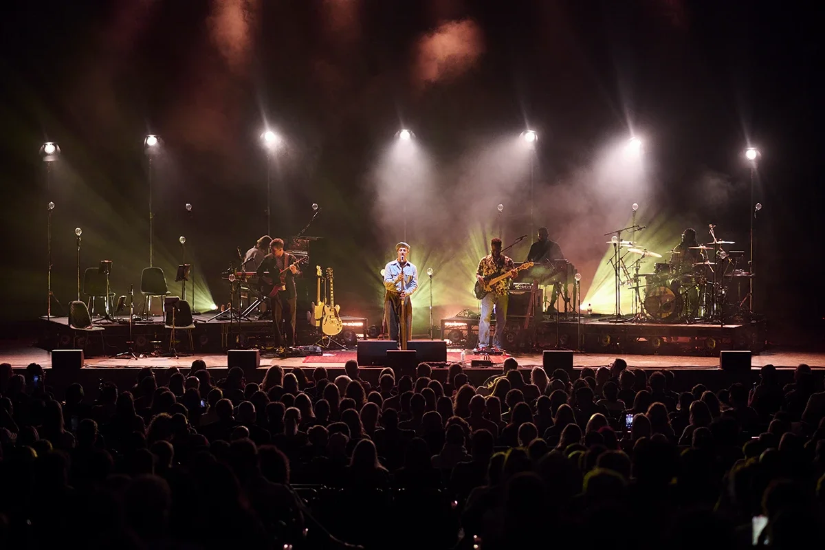 A band performs on stage in front of a large audience with bright stage lights and fog effects.