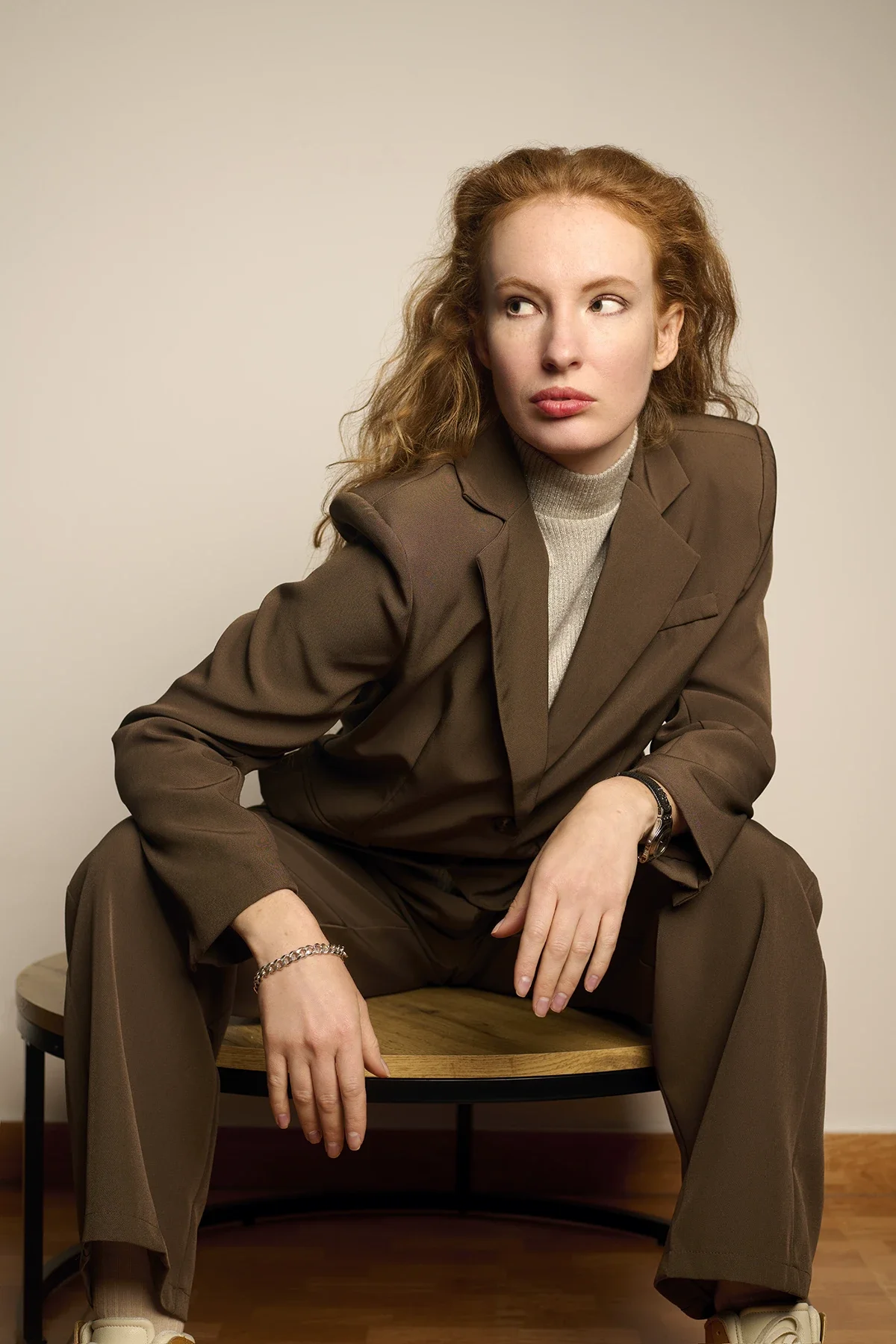 A woman with curly red hair sitting on a small round wooden table, wearing a brown suit and a beige turtleneck, gazing to her right with a serious expression.