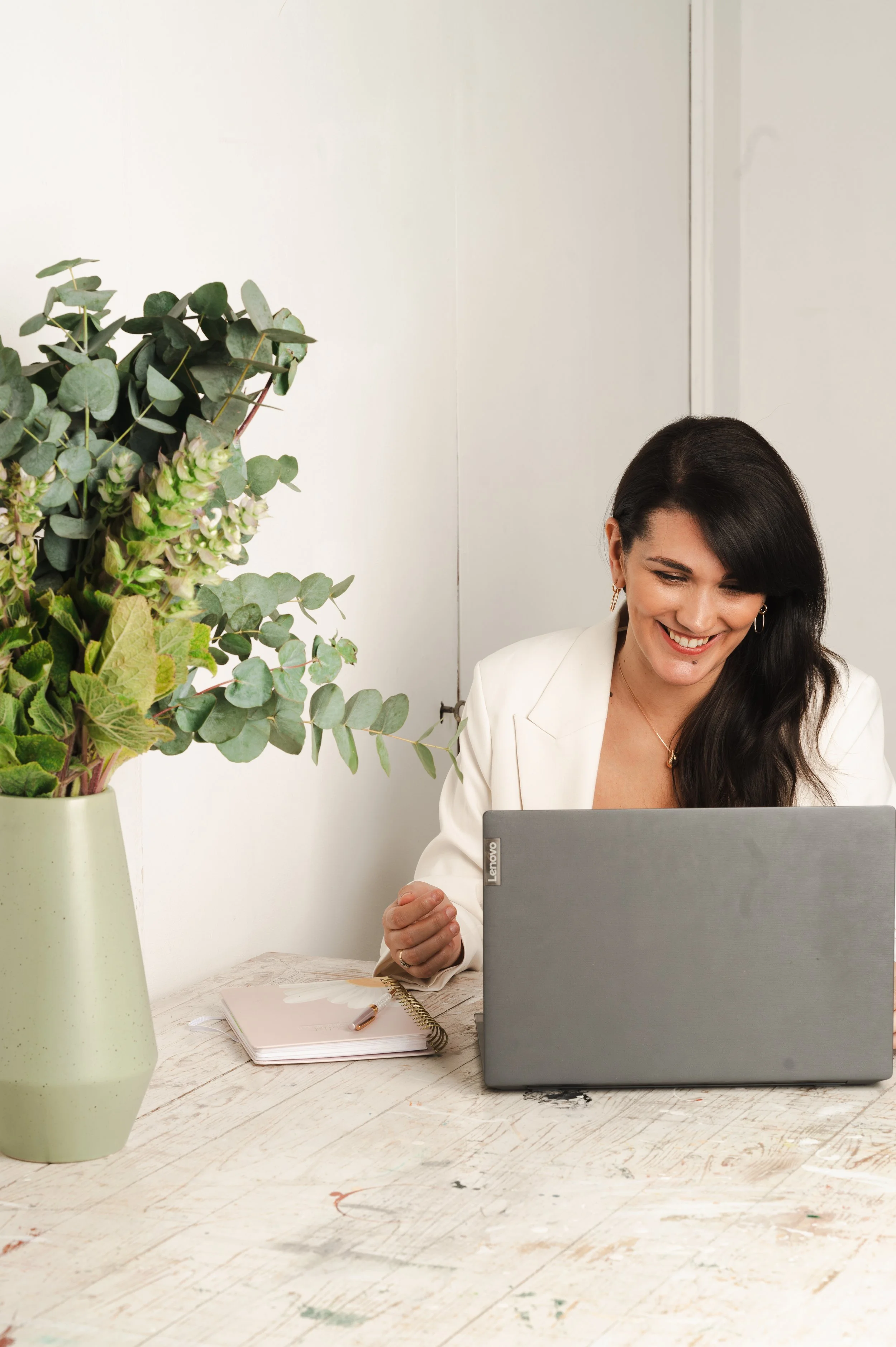 Isabel Fonseca, female leadership career coach in Amsterdam, and a white blazer smiling at her laptop while sitting at a desk with a large green plant and a notebook.