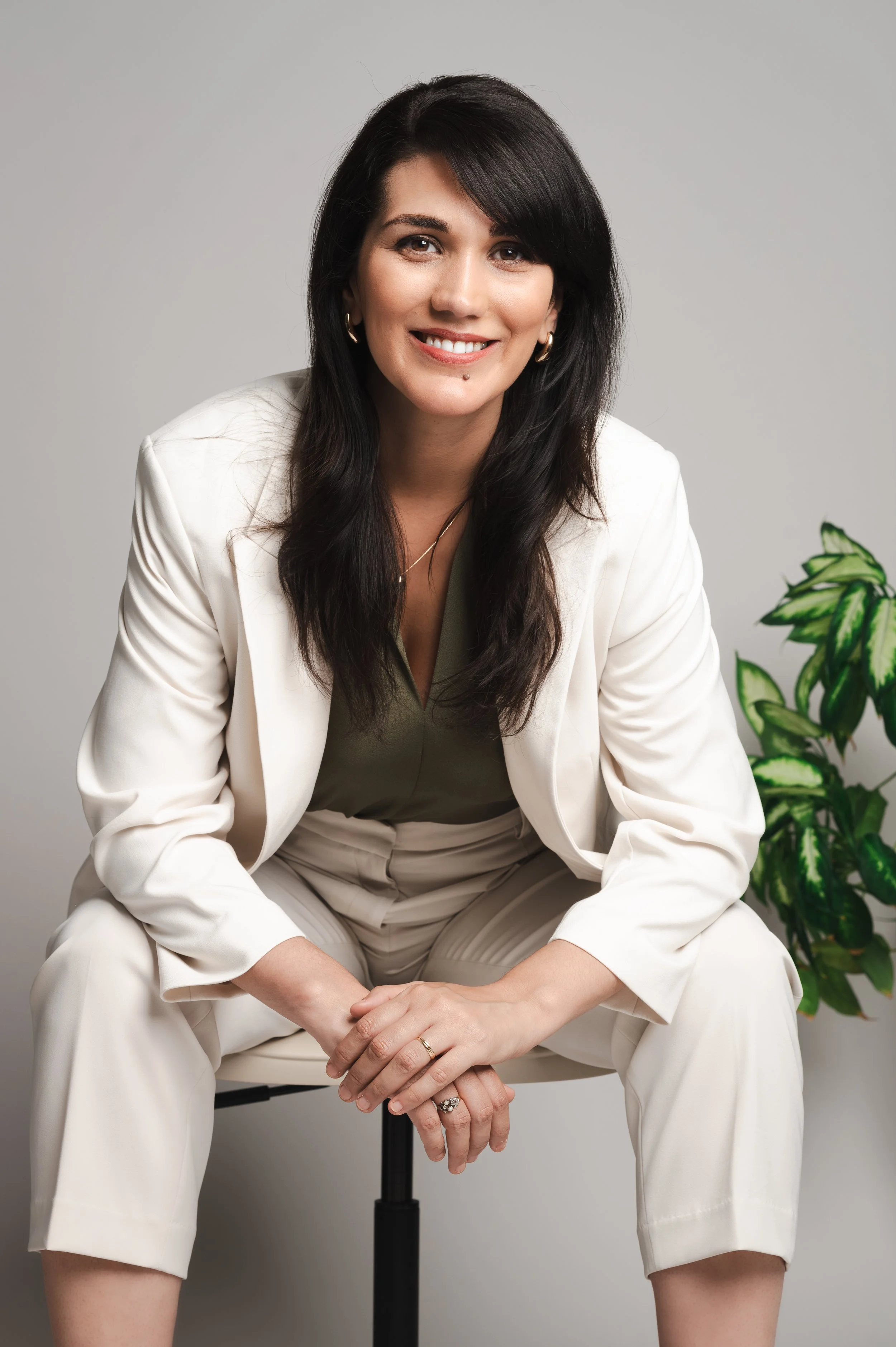 Isabel Fonseca, female leadership career coach in Amsterdam, smiling, wearing a light-colored suit, sitting on a chair with her hands clasped, with a potted plant to her right, against a neutral gray background.
