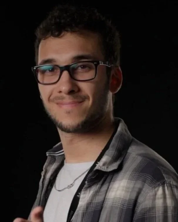 A young man with glasses, short curly hair, and a slight beard smiling, wearing a plaid shirt over a white t-shirt, against a black background.