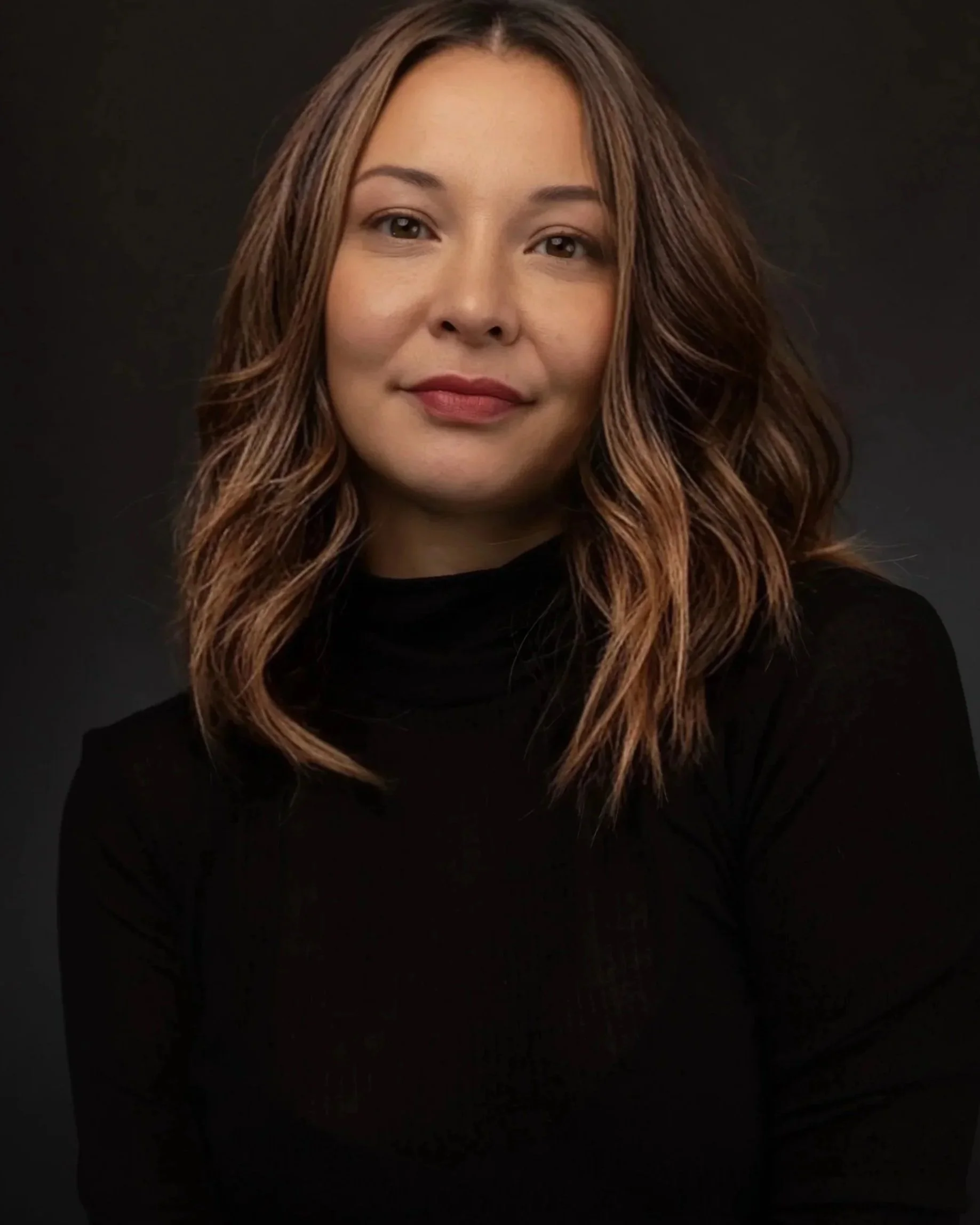 Portrait of a woman with wavy, shoulder-length hair, wearing a black top, against a dark background.
