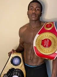 A shirtless young man holding two championship belts, standing against a plain wall.