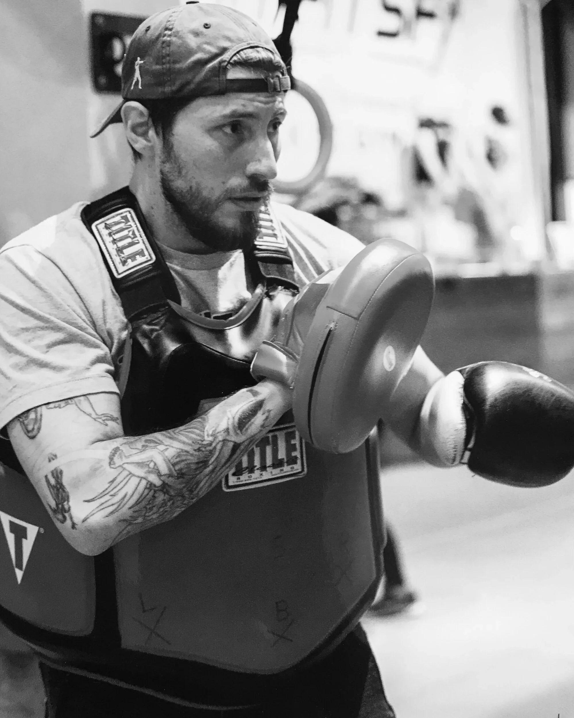 A man with tattoos on his arm, wearing a baseball cap backwards, and a T-shirt with patches on the shoulders, is holding an electric kettle or coffee pot.