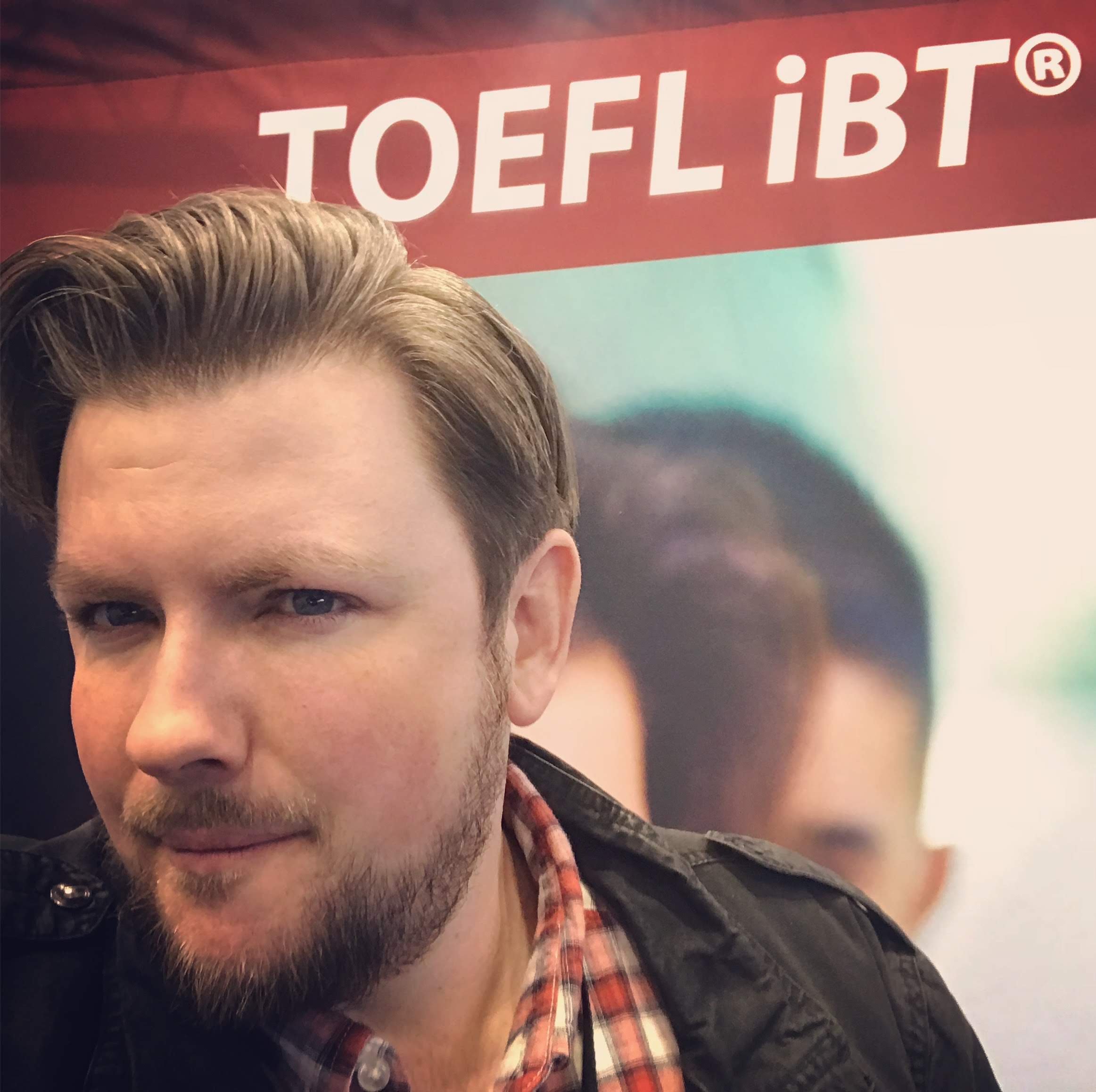 A young man with a beard and styled hair taking a selfie in front of a colorful background with the text 'TOEFL IBT' at the top.