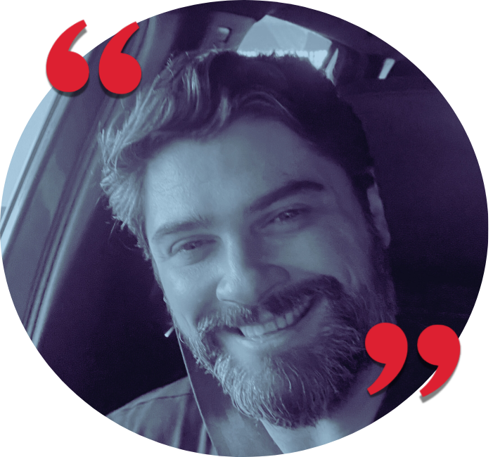 A smiling man with a beard and wavy hair, sitting in a vehicle, looking at the camera.