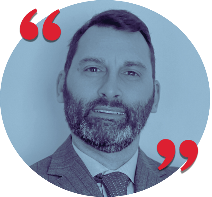 Portrait of a man with a beard wearing a suit and tie, smiling in front of a light-colored background with red quotation mark graphics.