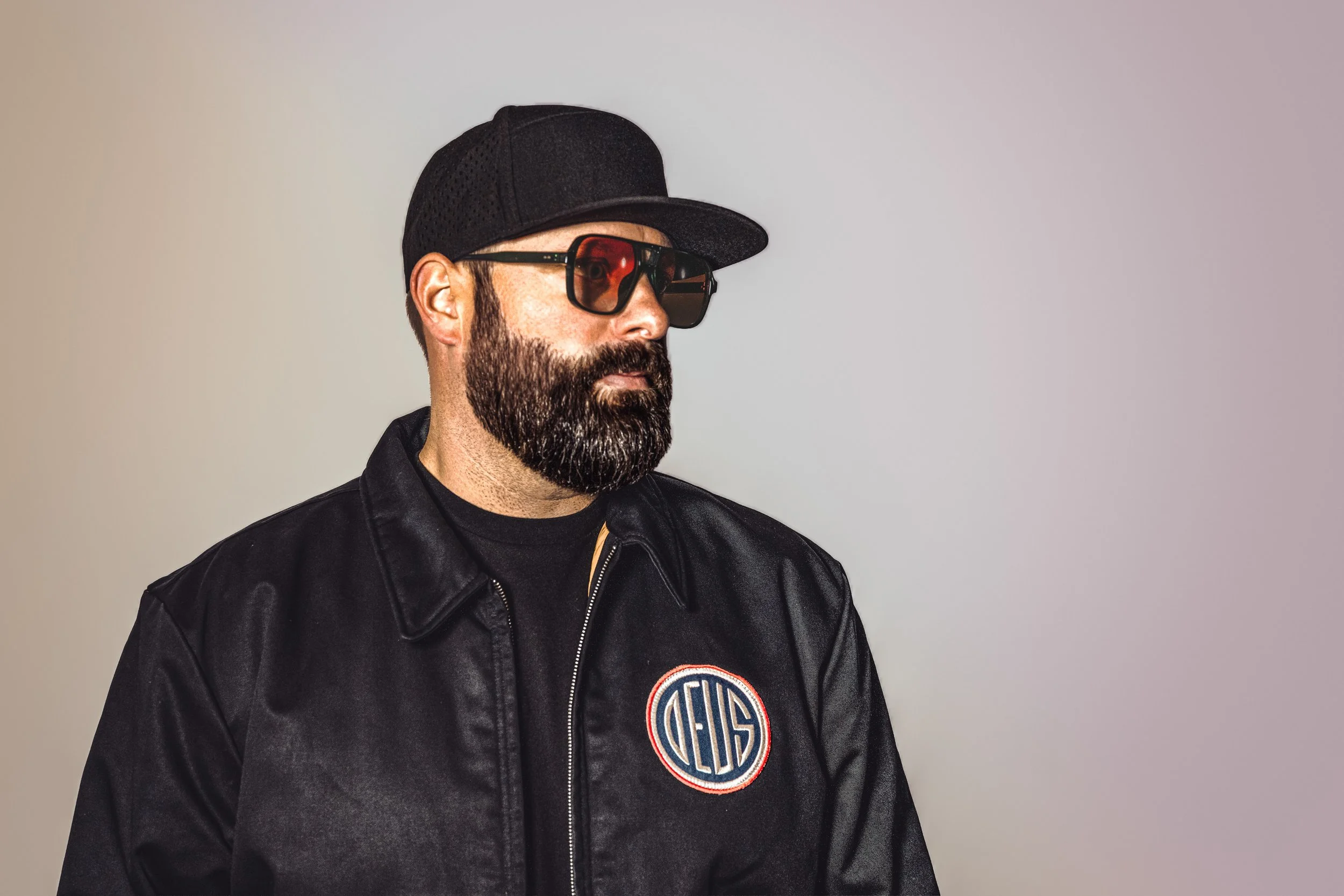Portrait of Jon, DJ and producer performing as Shares, wearing a black cap, tinted sunglasses, and a black jacket, against a neutral backdrop.