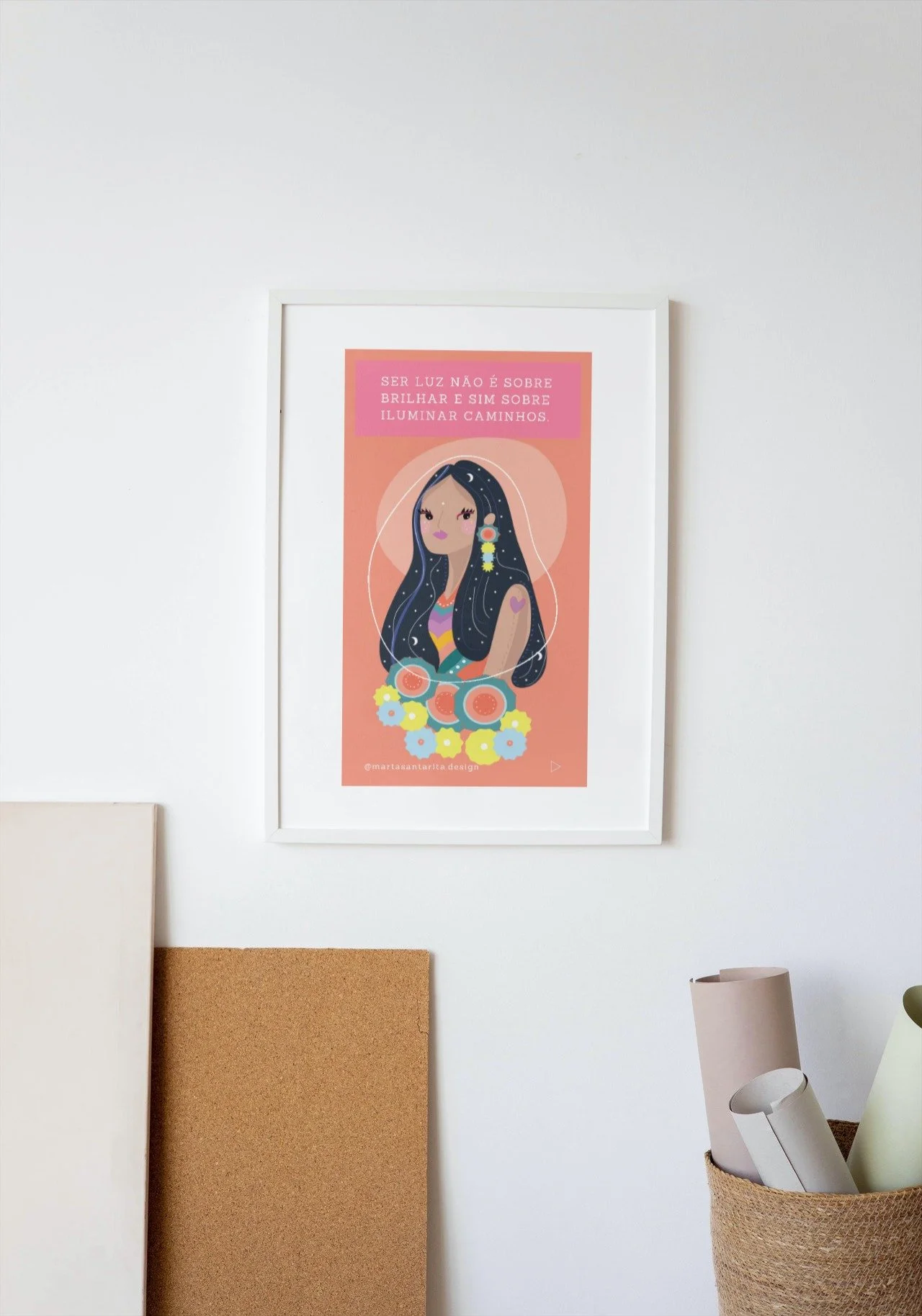 A wall with a framed artwork featuring an illustrated woman with floral patterns and the text in Portuguese. Below the frame are rolled papers in a basket and a corkboard leaning against the wall.