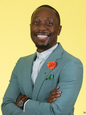 A man with dark skin in a light blue blazer, white button-up shirt, with a flower lapel pin, against a yellow background.