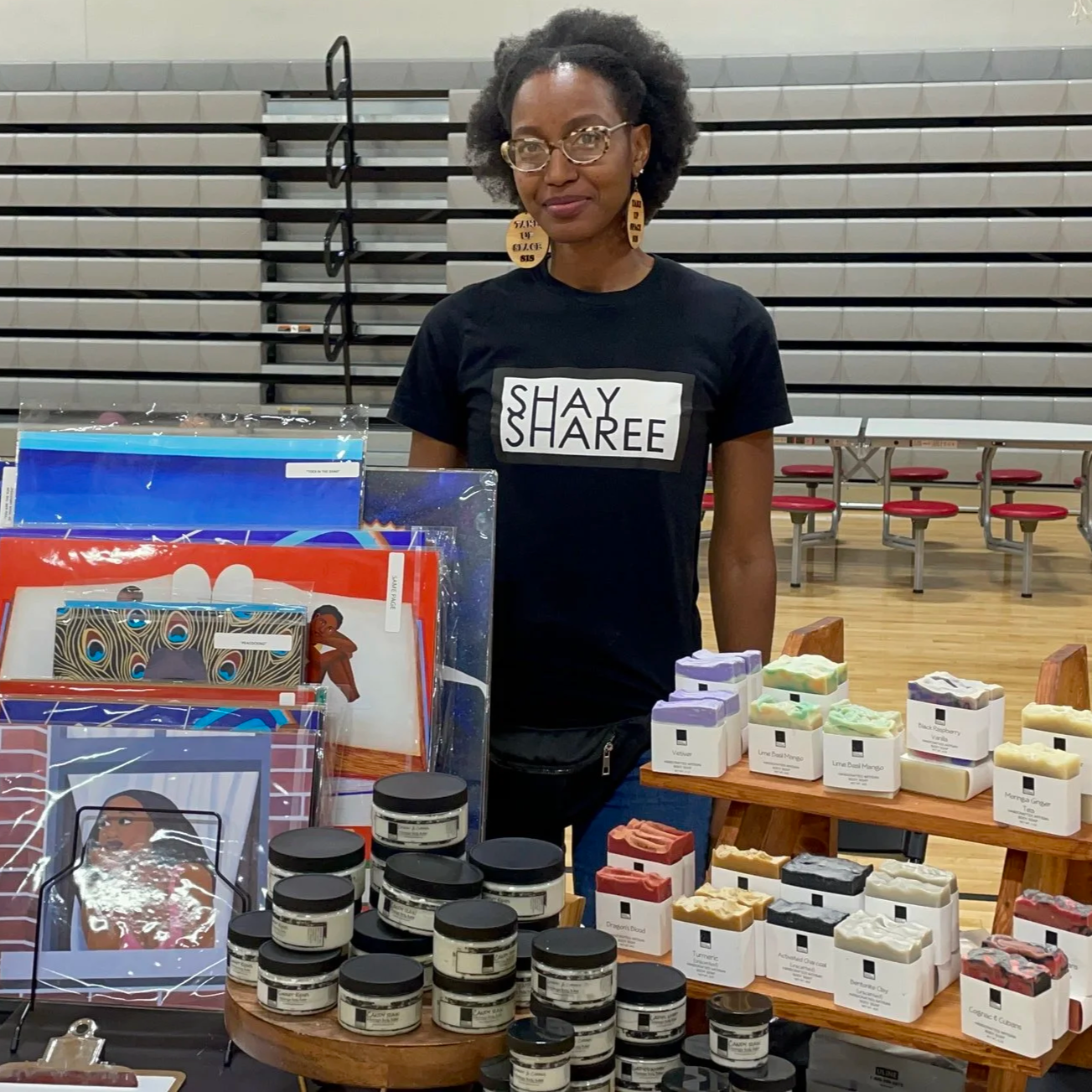 Woman standing behind a table with handmade soaps and skincare products at an indoor event, wearing glasses and a black T-shirt with 'SHAY SHAREE' printed on it.