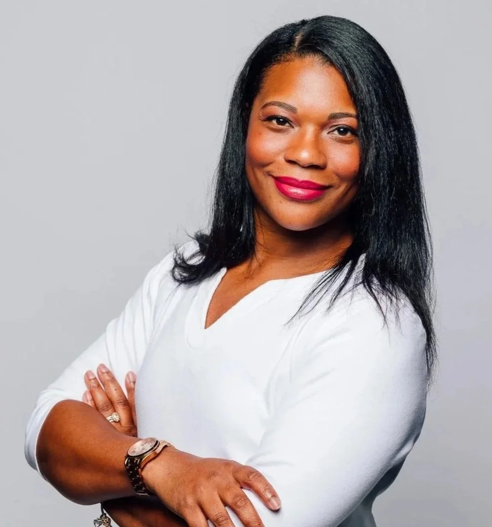 A woman with straight black hair, wearing a white top, smiling with arms crossed against a neutral background.