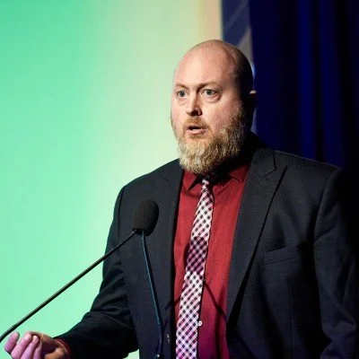 A man with a beard in a suit speaking at a podium with a microphone.