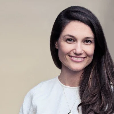 A woman with long dark hair smiling and wearing a white top, posing against a plain background.