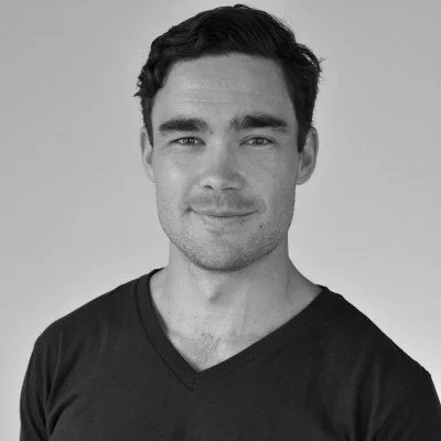Black and white portrait of a young man with dark hair, wearing a V-neck T-shirt, smiling slightly, against a plain background.