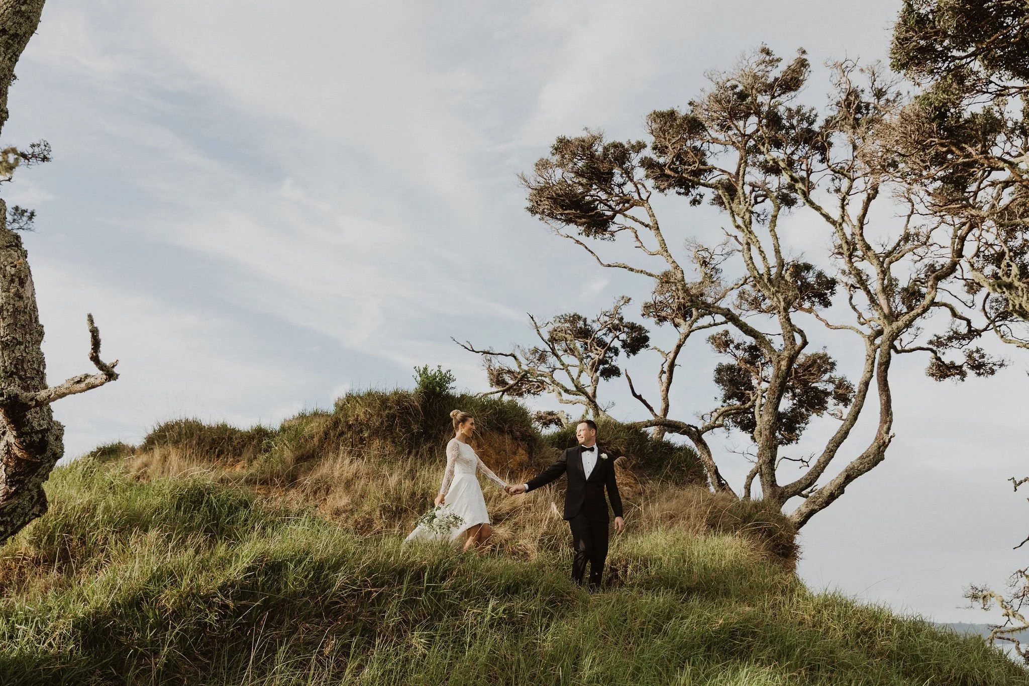 Bride and groom holding hands, walking on grass with trees in the background at Bayly's Farm in Paihia.
