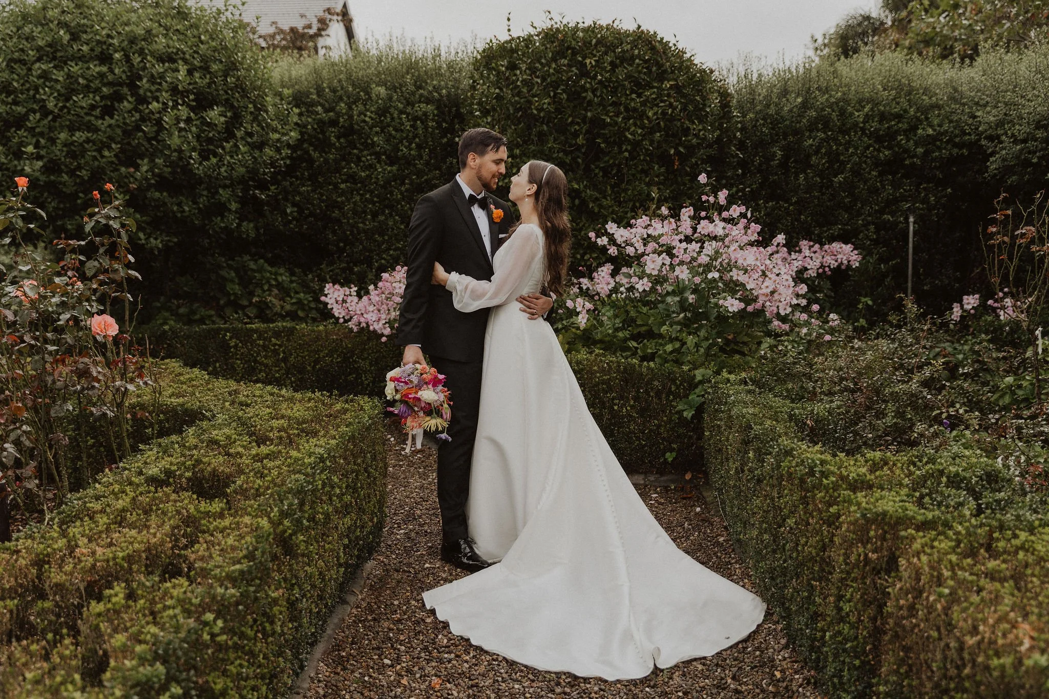 Bride and groom embracing in a garden, surrounded by hedges and pink flowers, holding a colorful bouquet in Cambridge at Henley Hotel.