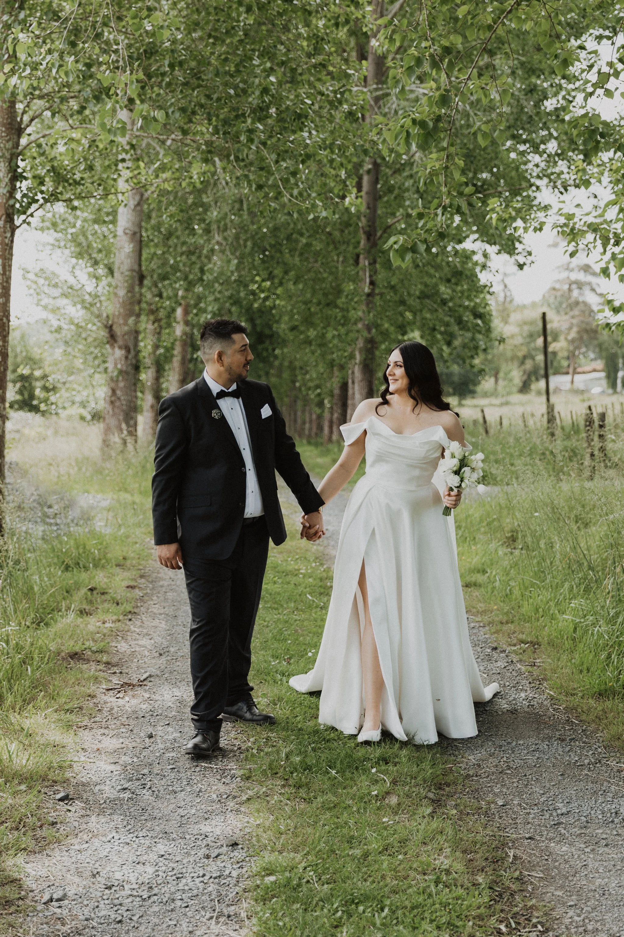 A bride and groom walking hand-in-hand on a path lined with trees, outdoors in a natural setting in Auckland. The bride is wearing a white wedding dress and holding a bouquet, while the groom is dressed in a black suit Auckland wedding photography