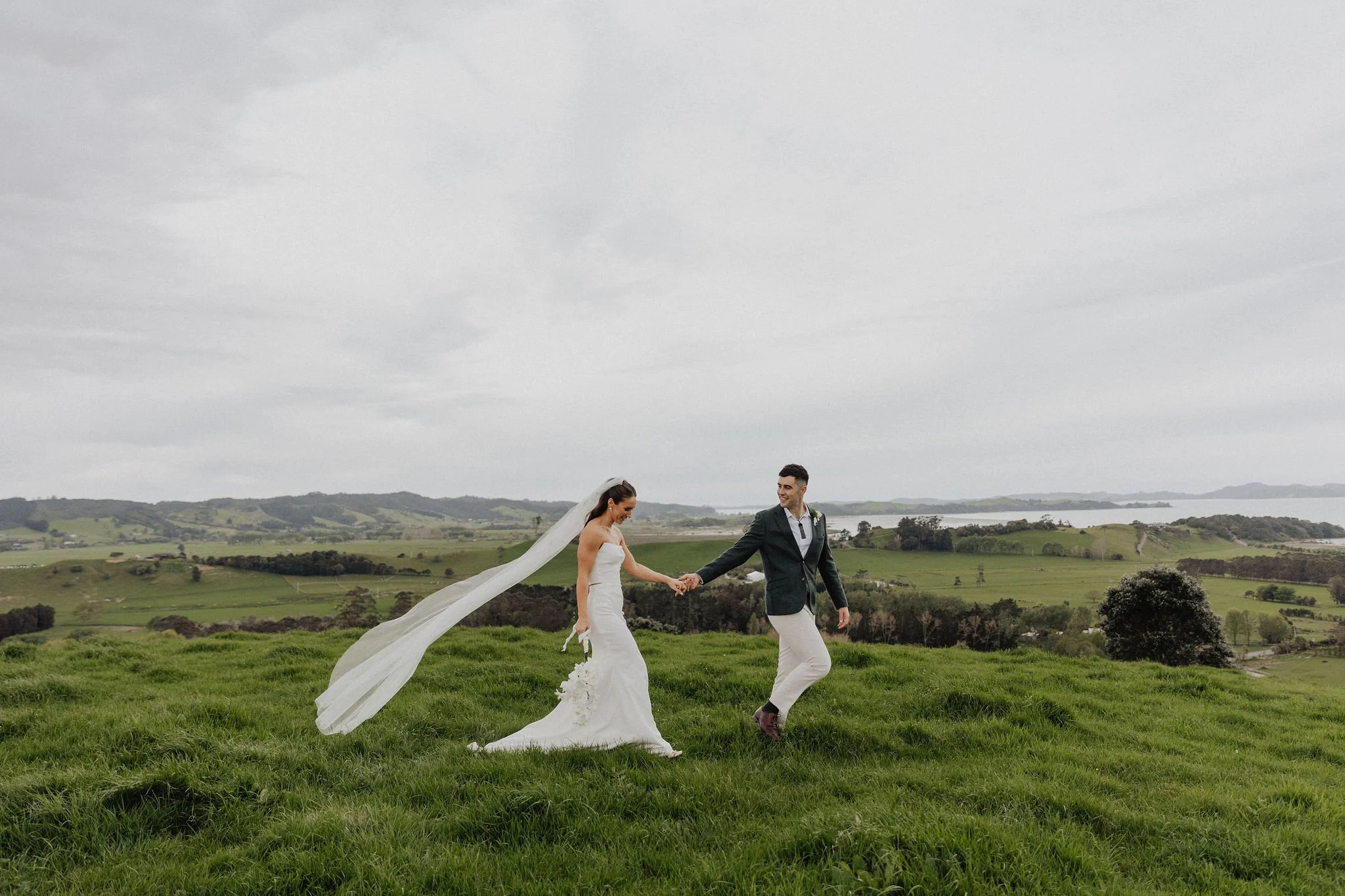 Bride and groom walking hand in hand on grassy hilltop, scenic landscape in background at Kauri Bay Boomrock, Auckland