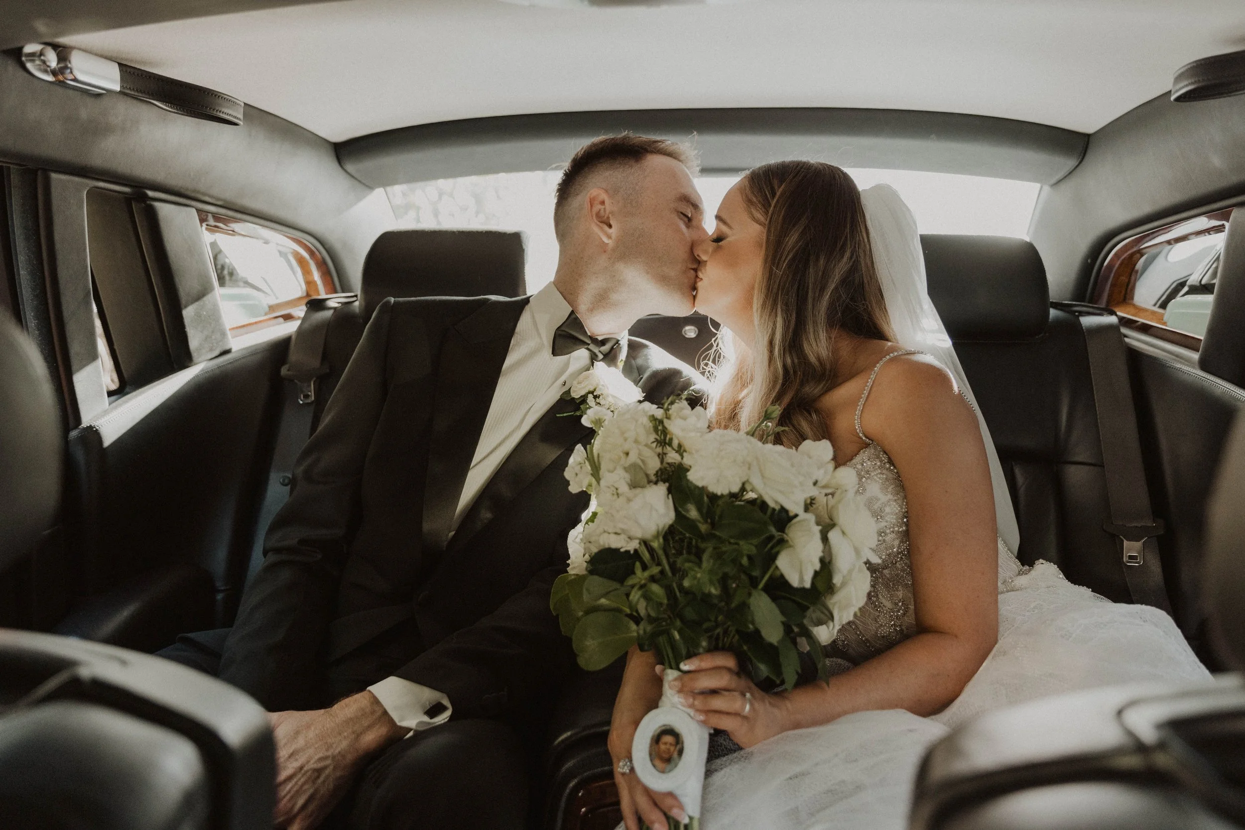 Bride and groom kissing in the back of a car, holding a bouquet of white flowers at a luxury Auckland wedding.