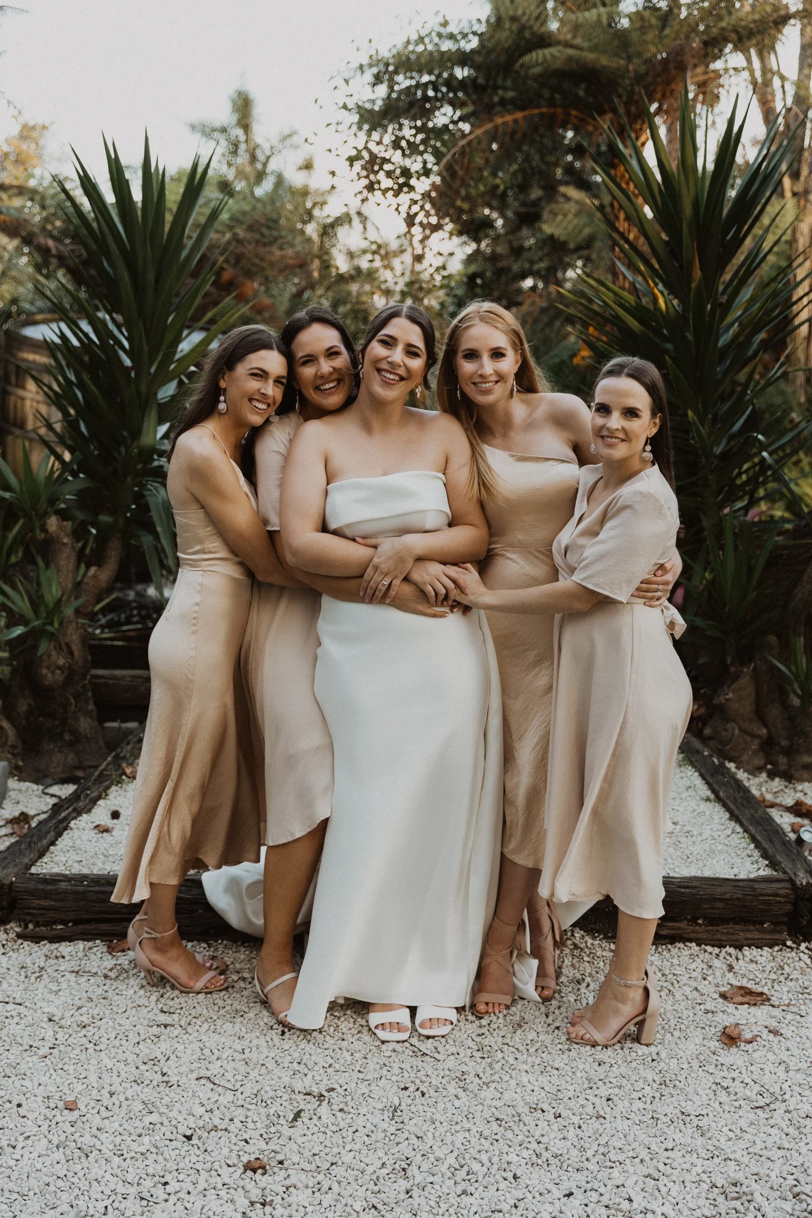 Bridal party with bride and bridesmaids posing happily outdoors in neutral dresses at Markovina in Auckland.