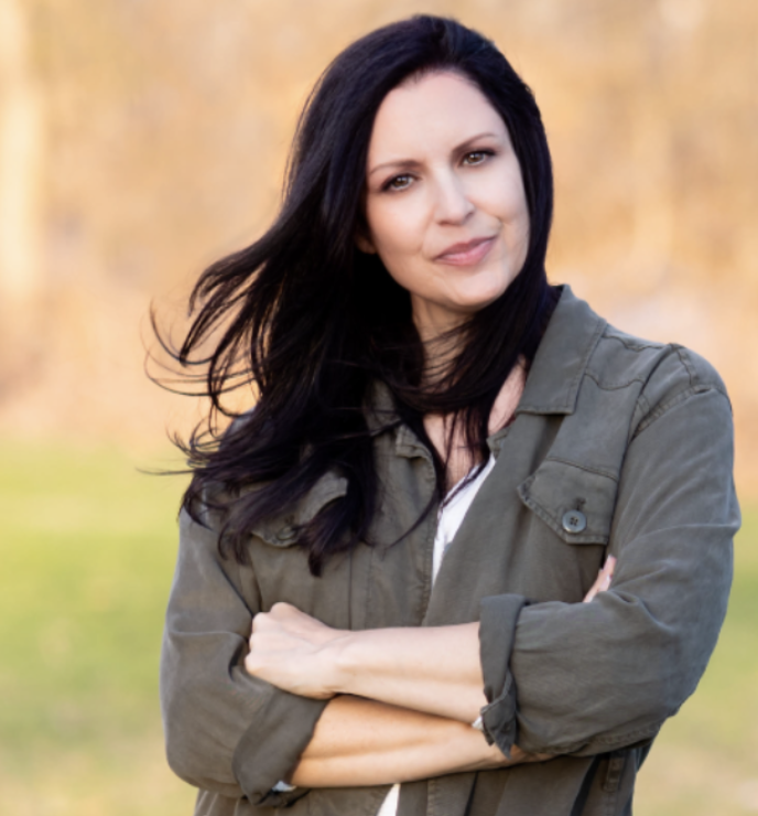 A woman with long dark hair standing outdoors with arms crossed, wearing an olive green jacket, in front of blurred fall-colored trees.