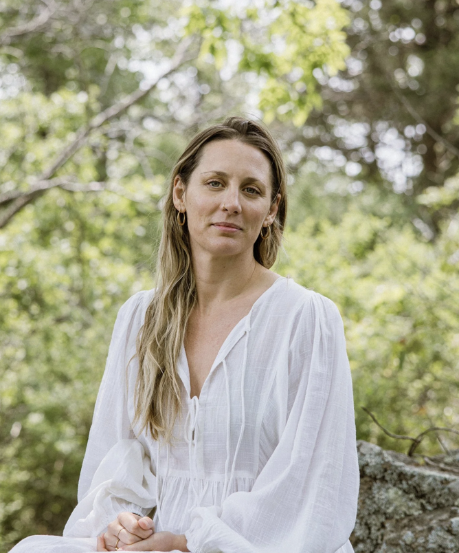 A woman with long blonde hair wearing a white blouse sitting outdoors with trees and greenery in the background.