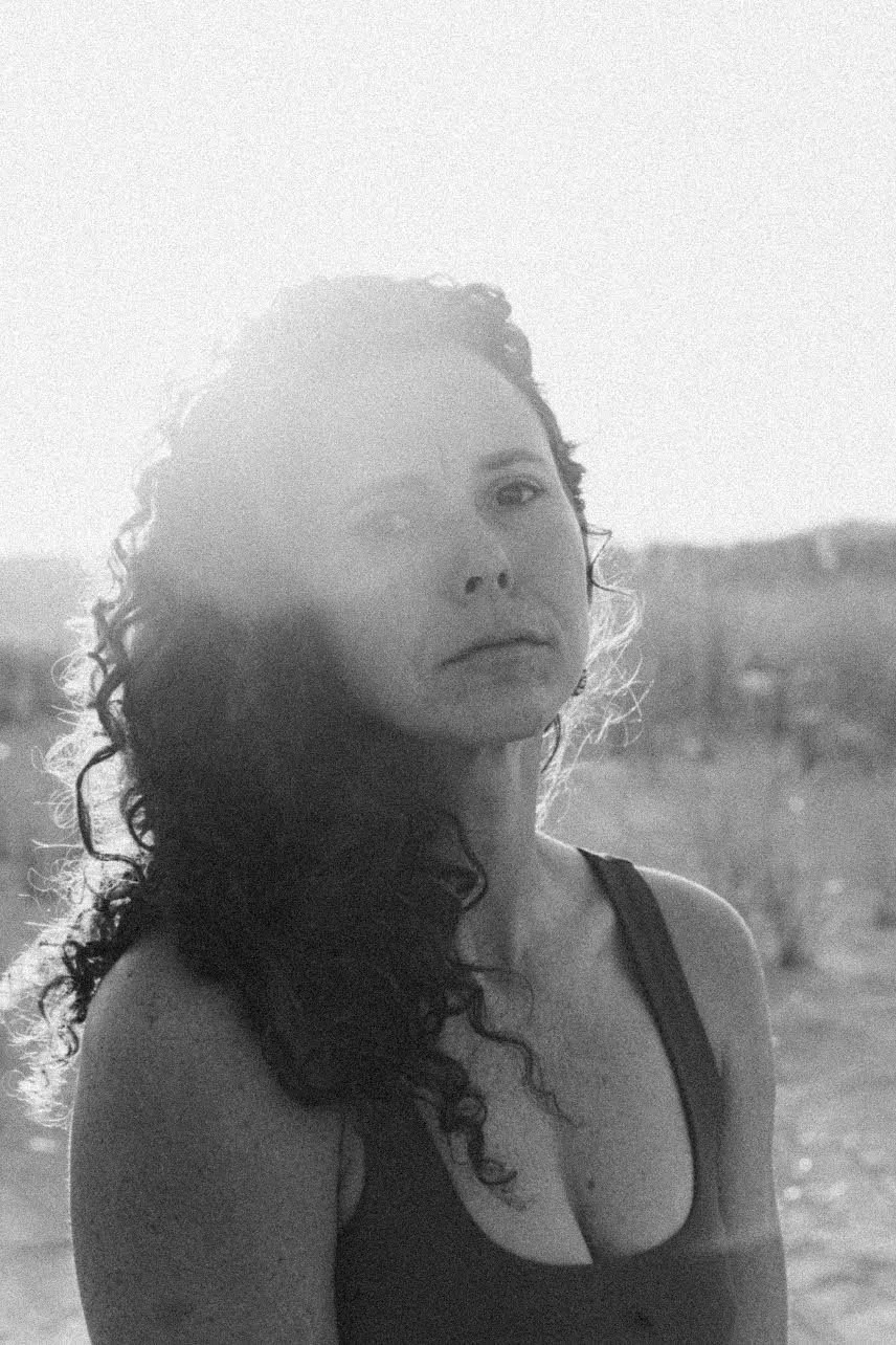 Black and white photo of a young woman with curly hair looking at the camera, outdoors with blurred landscape in the background.