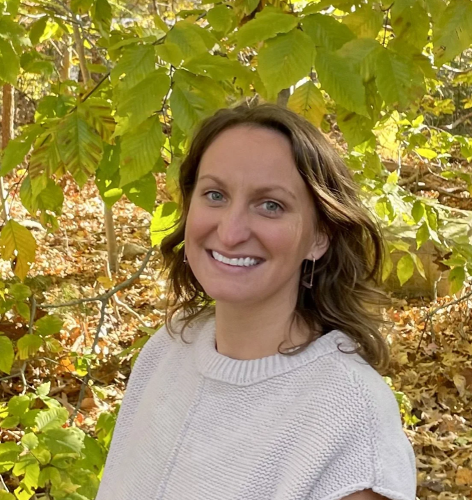 A woman with wavy brown hair smiling outdoors amidst green and autumn leaves.