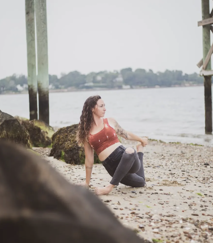 A woman with curly hair and tattoos sitting on a rocky beach near water, wearing a red crop top and black pants.